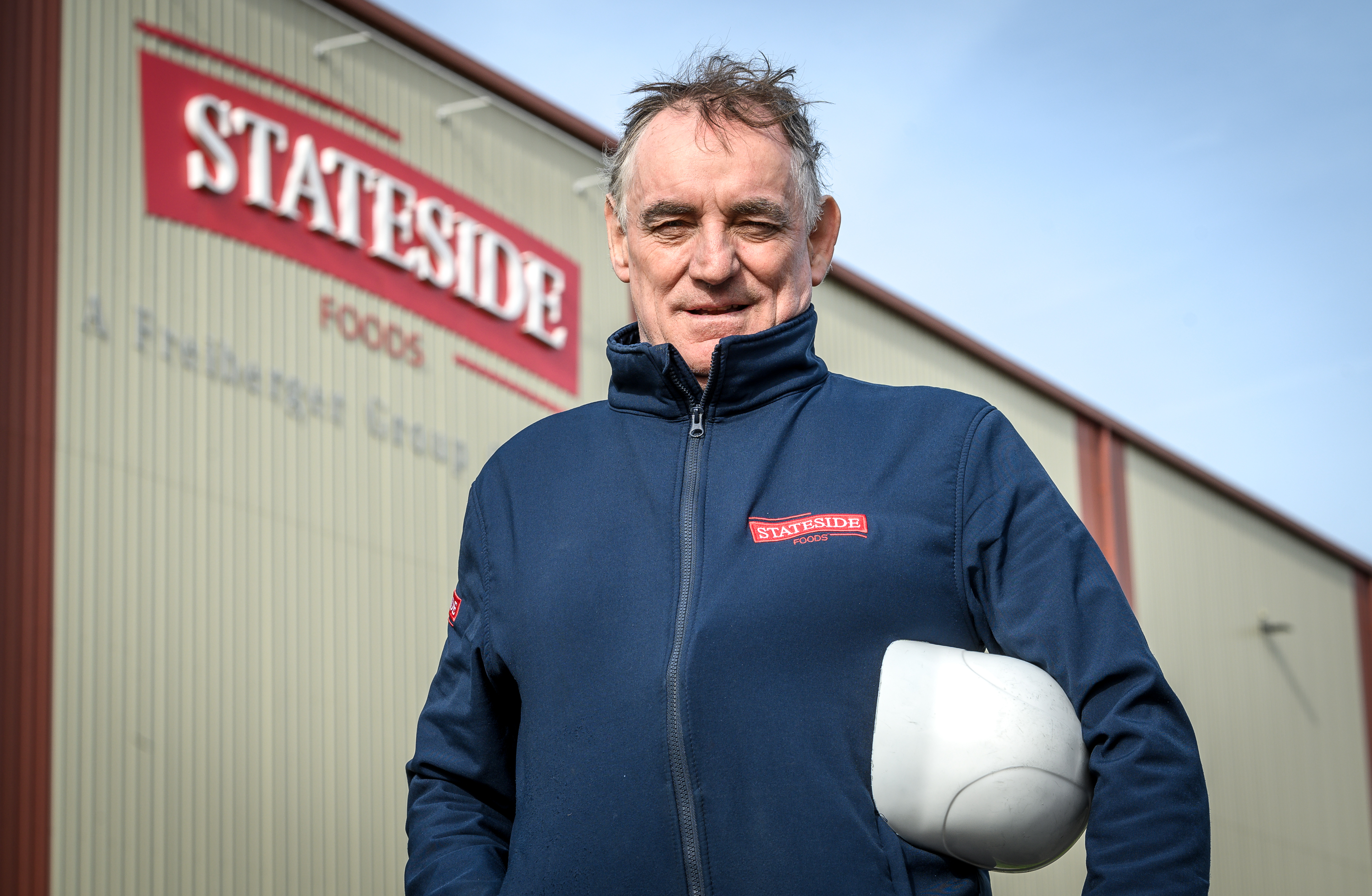 A workshop customer stands in-front of a 
"Stateside Foods" warehouse, wearing a blue zip-up, branded jumper and holding a white hardhat.